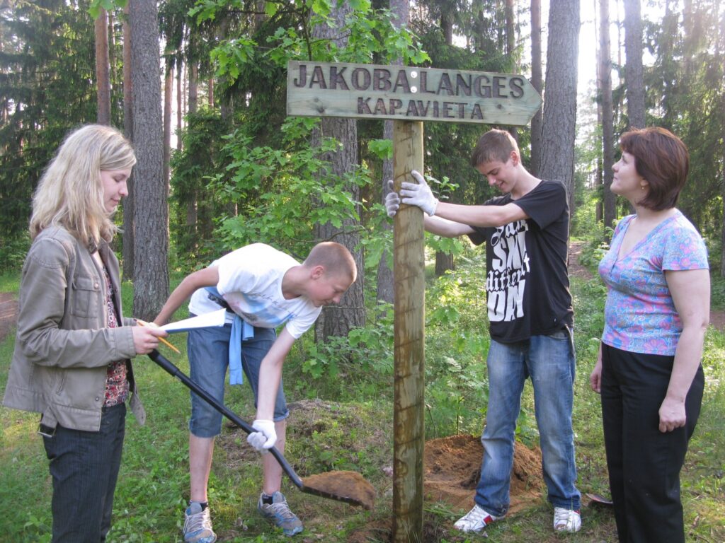 Skolnieki Jānis Lauris (no kreisās) un Kārlis Ērģelis vasaras brīvlaikā atnāca palīgā savai audzinātājai Janai Vīksnai (no labās) un pašvaldības ainavu arhitektei Agijai Bērtiņai uzstādīt norādes uz mācītāja Jakoba Langes atdusas vietu. 
Autors: Sandra Pētersone