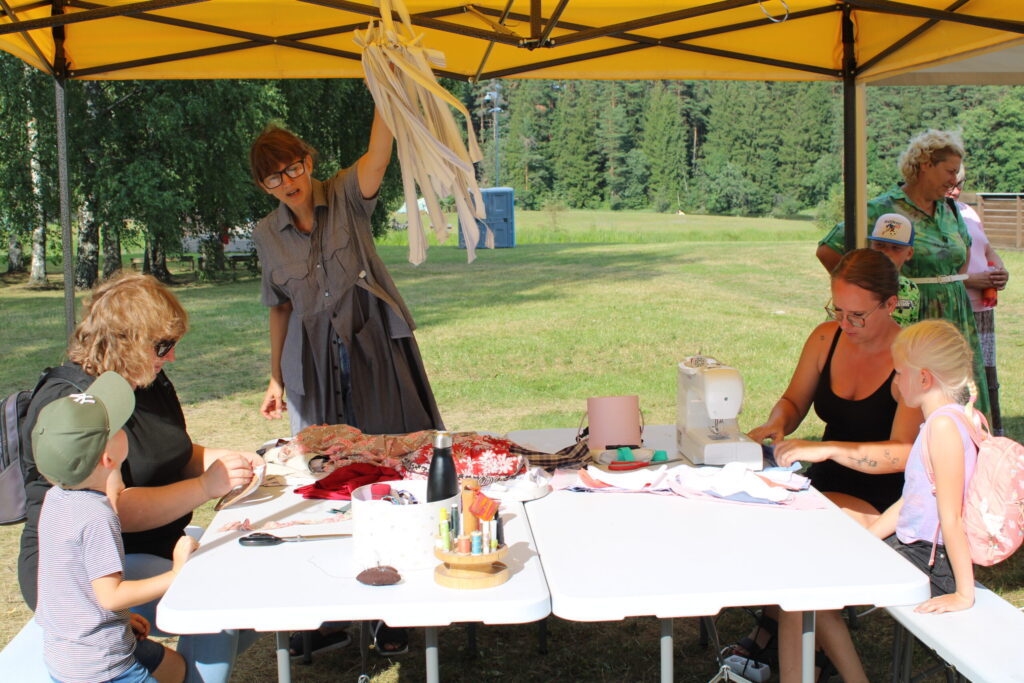 Variņiete Madara Kušnere (no labās) piedāvāja iesaistīties tekstila pārveides darbnīcā – šūt matu lentes un gumijas no lietotiem vīriešu krekliem, bet dizainere Aiva Zīle no Apes, apģērbu zīmola “Zīle” īpašniece (stāv kājās), – uzšūt karodziņu virteni vai dekoru no lentītēm.