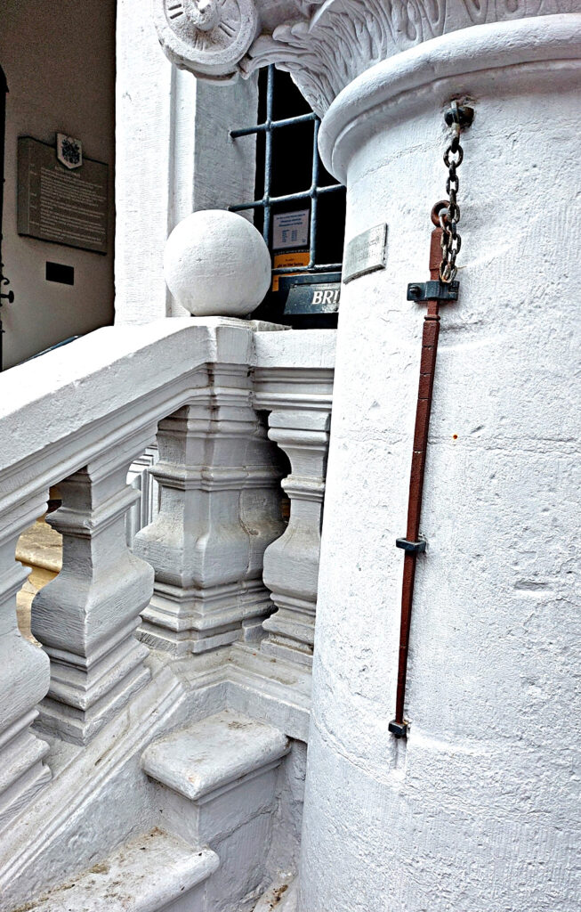 Pie rātsnama sienas ar ķēdi piestiprināts senais garuma etalons.