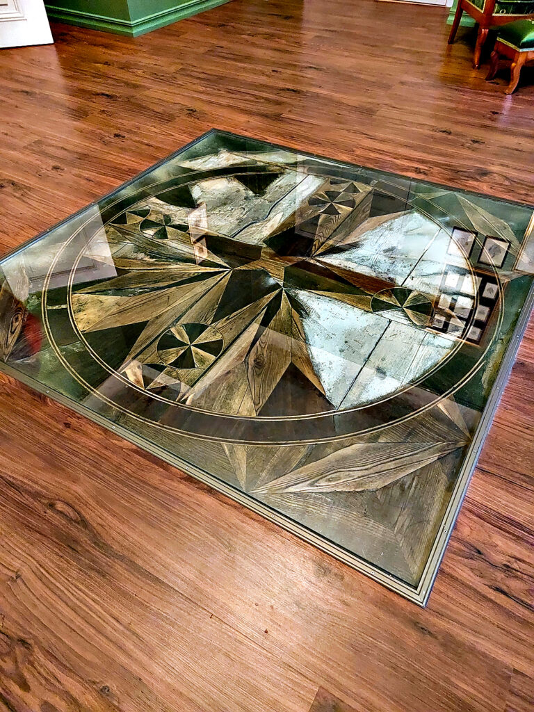 Oriģinālās grīdas fragments. Uz tā virsū ilgu laiku atradies ļoti smags rakstāmgalds, ko parasti nepārbīdīja. Kad to nocēla, apakšā atklājās oriģinālā grīda.