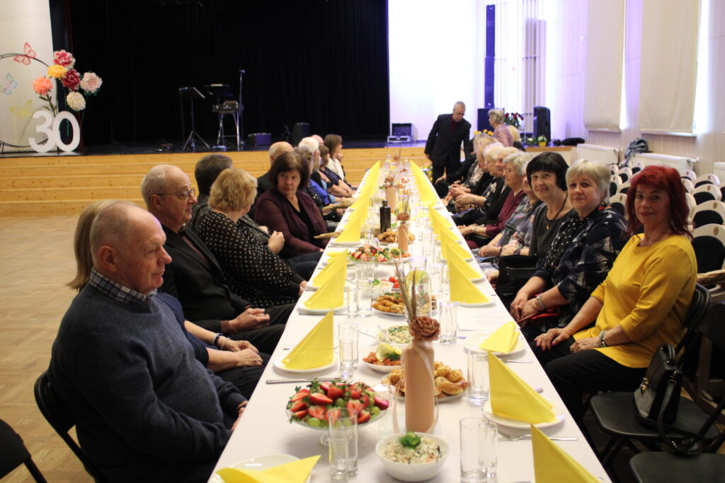 Jubilejas pasākums Smiltenes Kultūras centra lielajā zālē sapulcināja gandrīz 100 senioru no Smiltenes novada. Klātie galdi viņus gaidīja visapkārt zālei.
